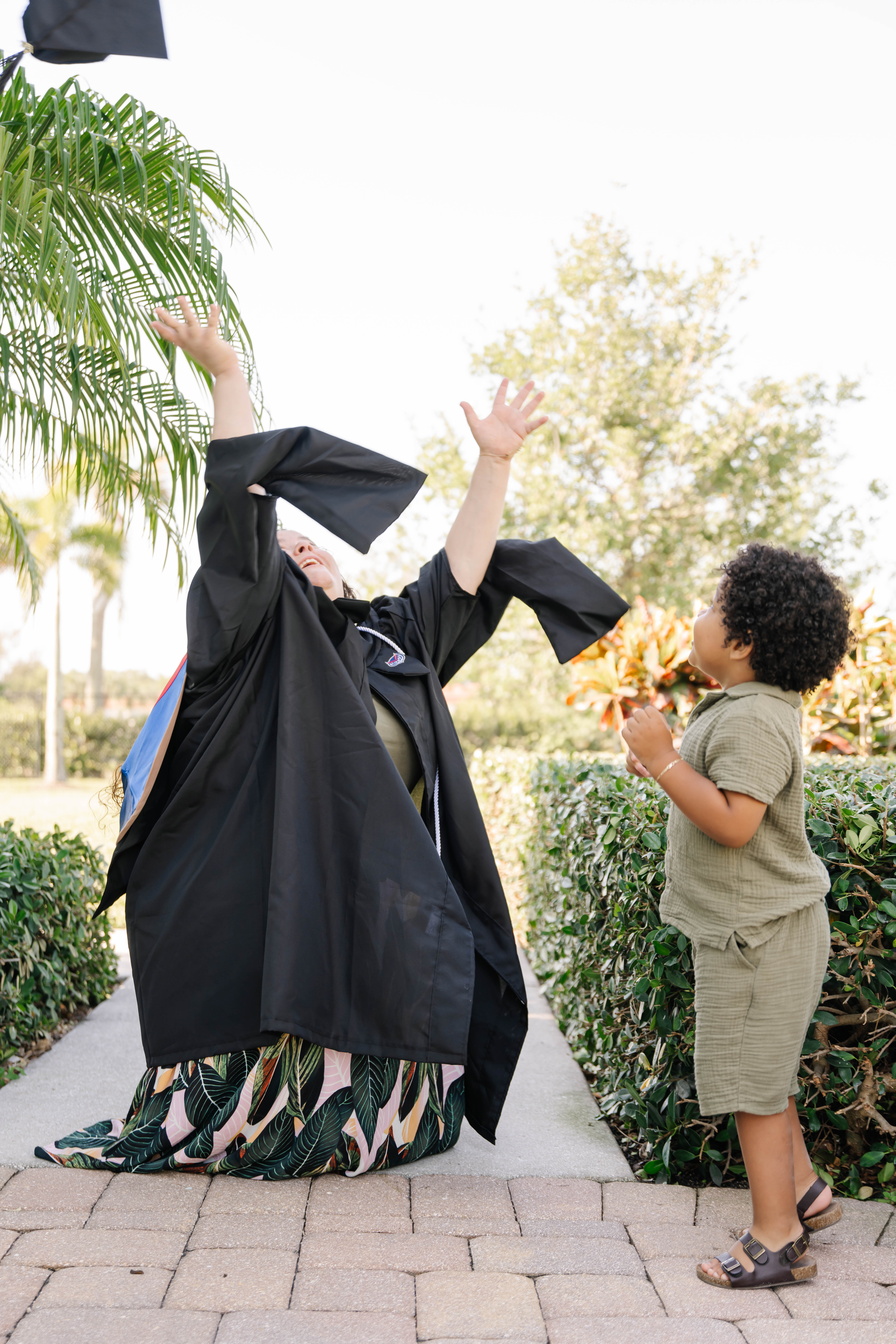 Woman in graduation gown throwing cap in air with young son beside her
