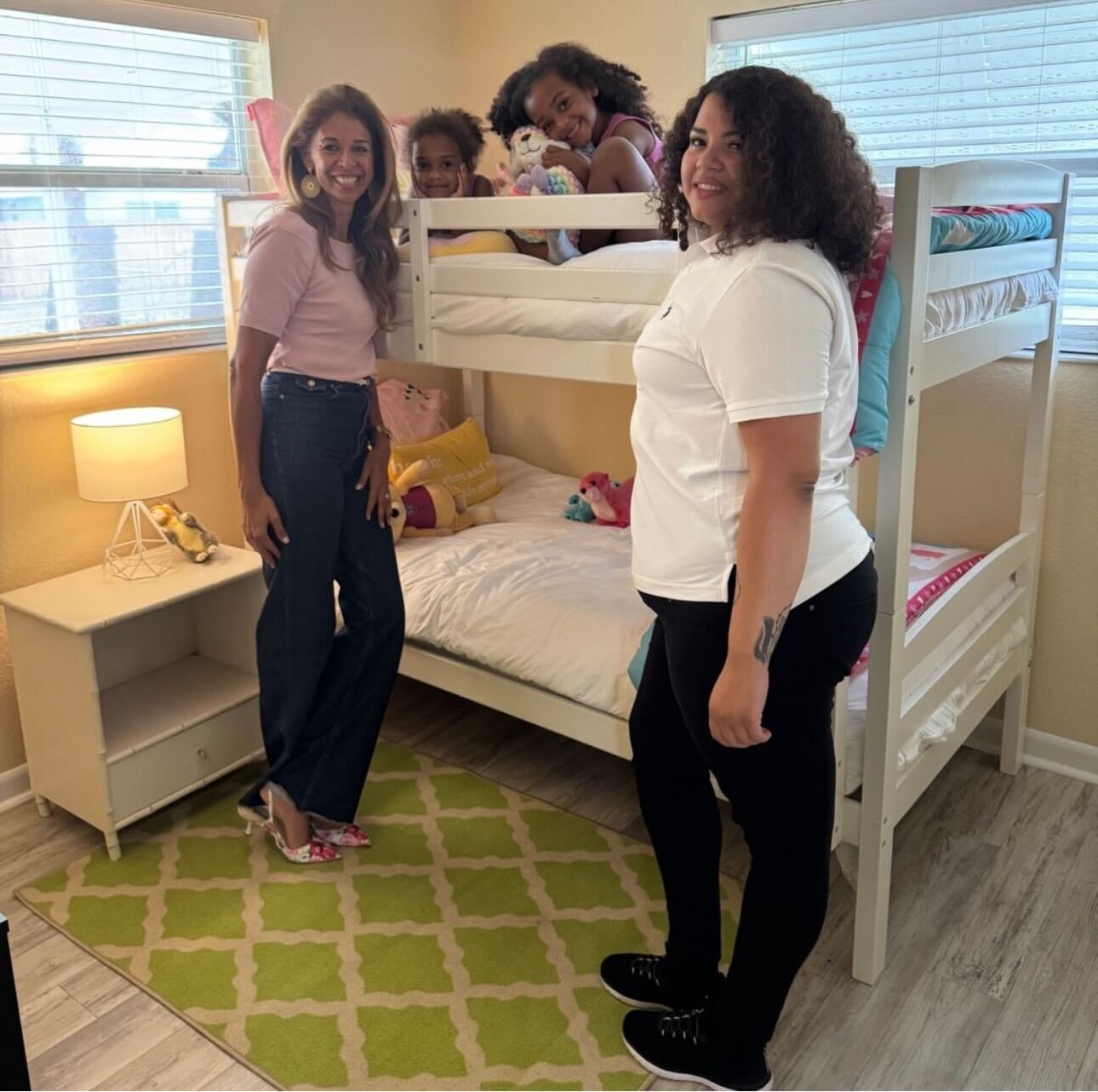 Two women standing next to a bunk bed with two children sitting on top.